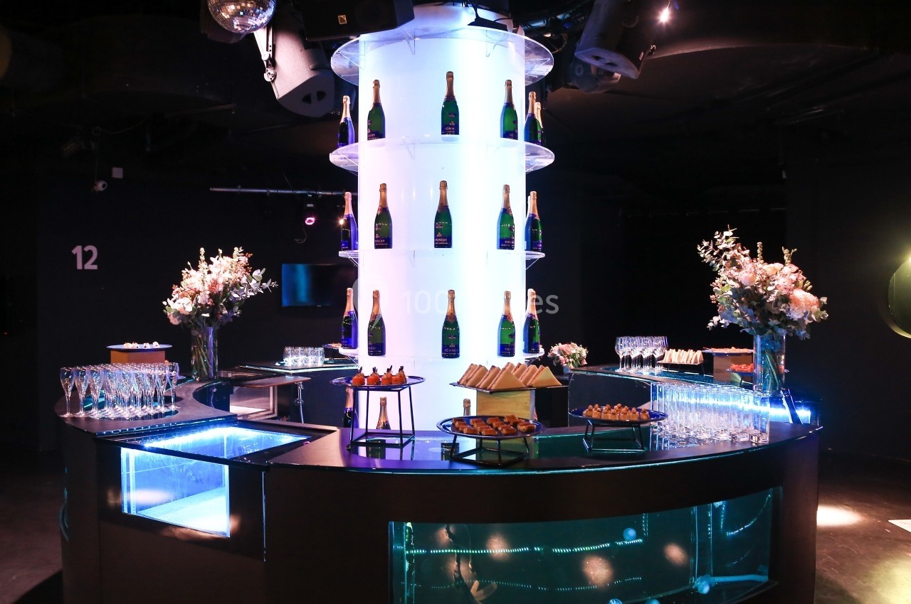 Bar circulaire illuminé avec bouteilles de champagne, verres, fleurs et amuse-bouches dans une ambiance sombre et élégante.