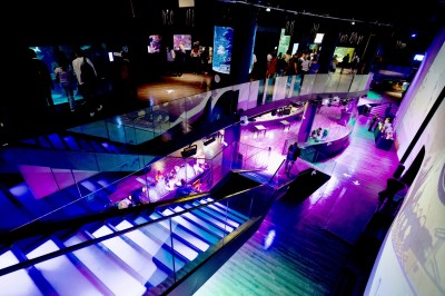 Tunnel d'aquarium avec un grand bassin à droite, éclairages colorés et écran lumineux sur le mur gauche.