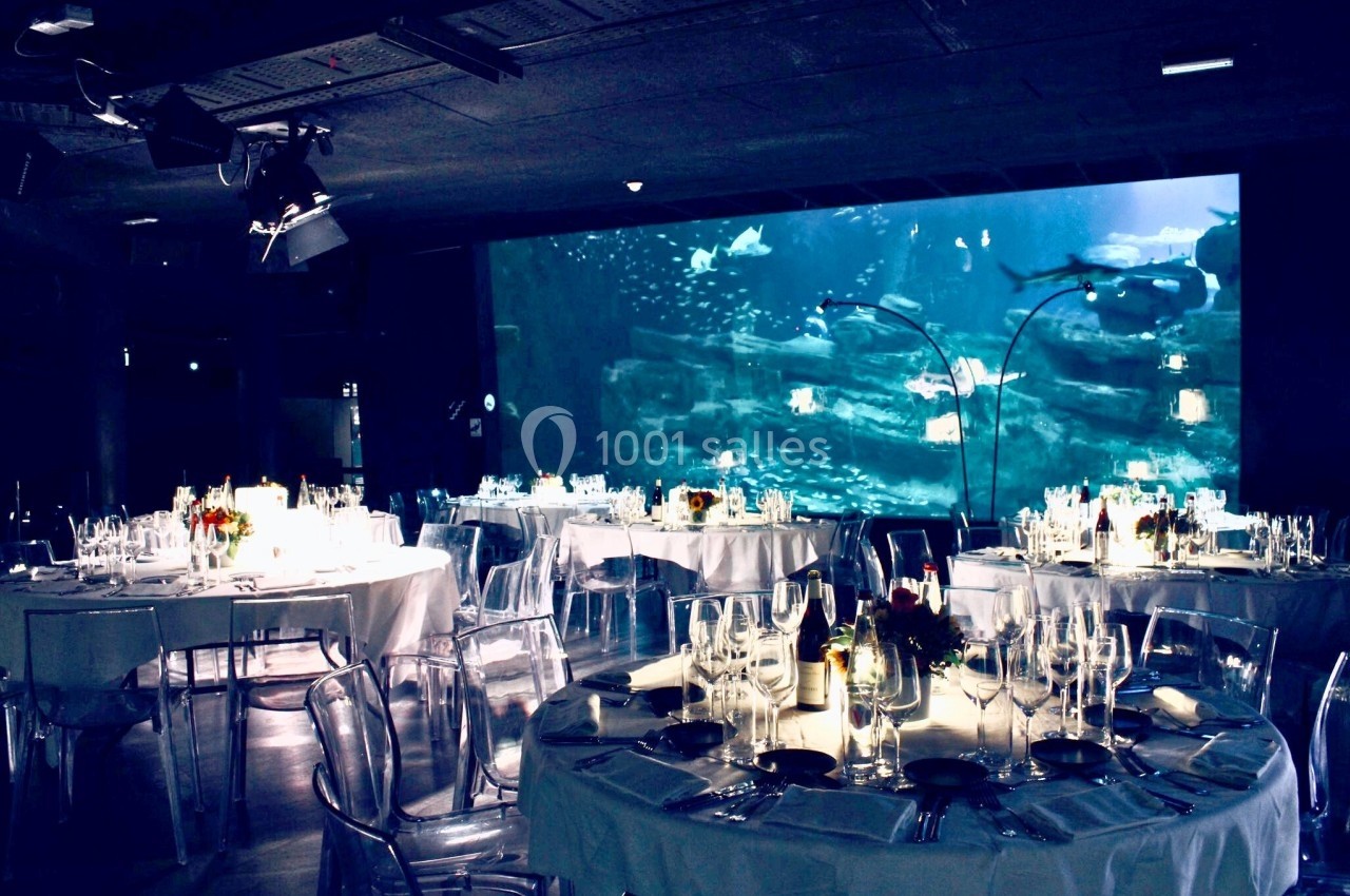 Salle de réception avec tables élégamment dressées, éclairage tamisé et grand aquarium en arrière-plan.