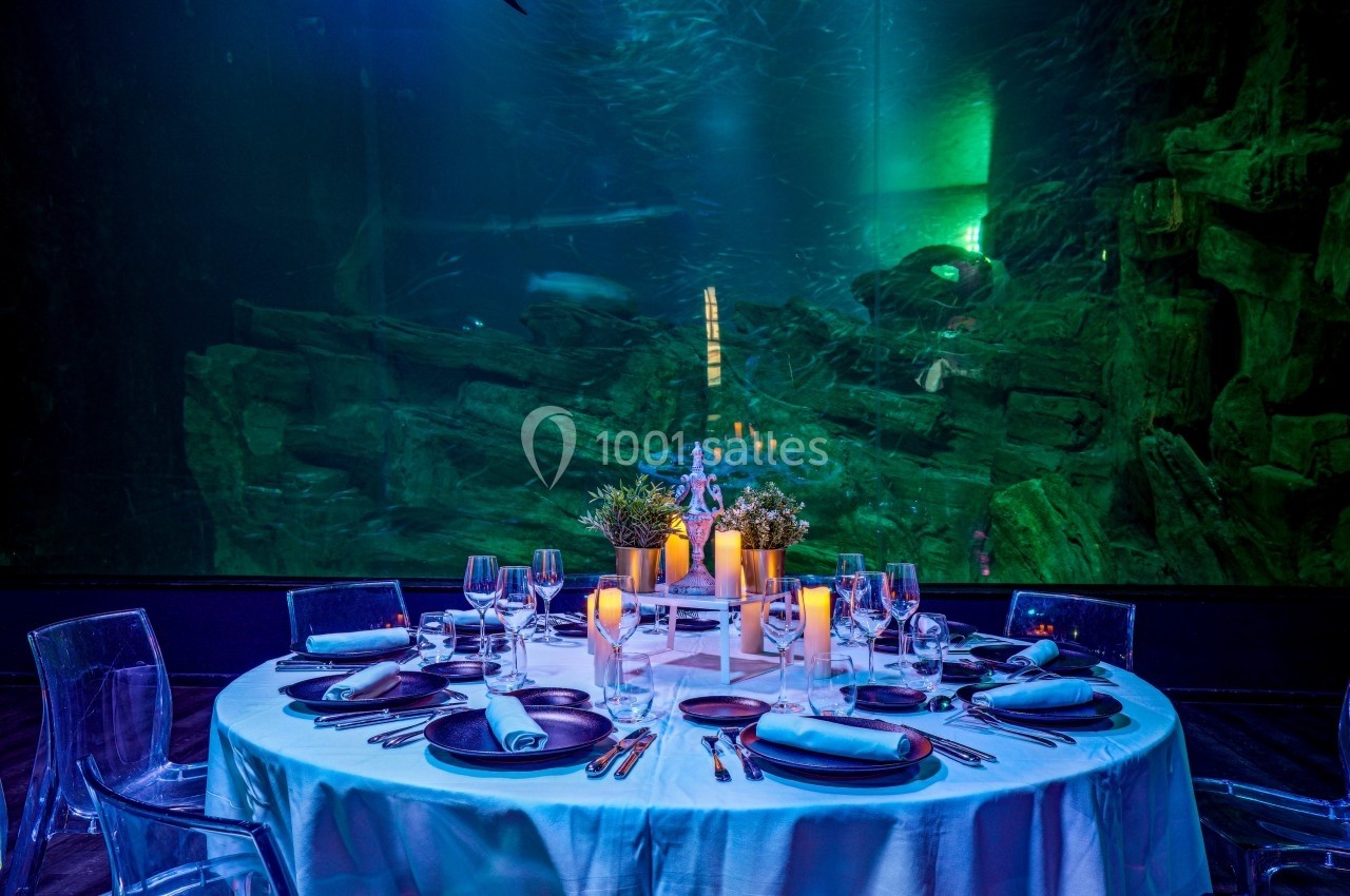 Table élégamment dressée avec des bougies et des plantes, placée devant un grand aquarium éclairé en bleu et vert.