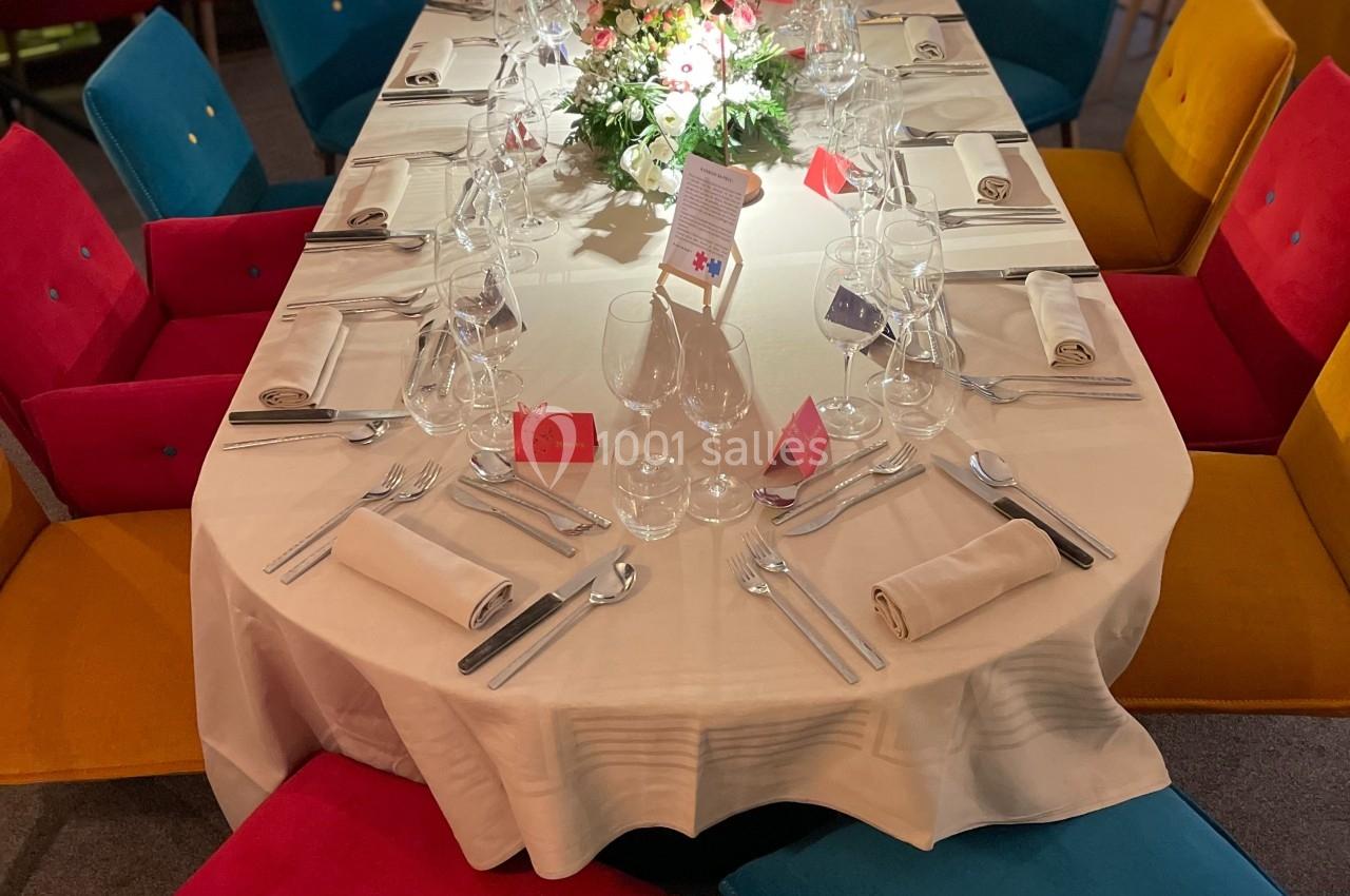 Table ovale dressée avec nappes blanches, verres, couverts et centre de table fleuri, entourée de chaises colorées.