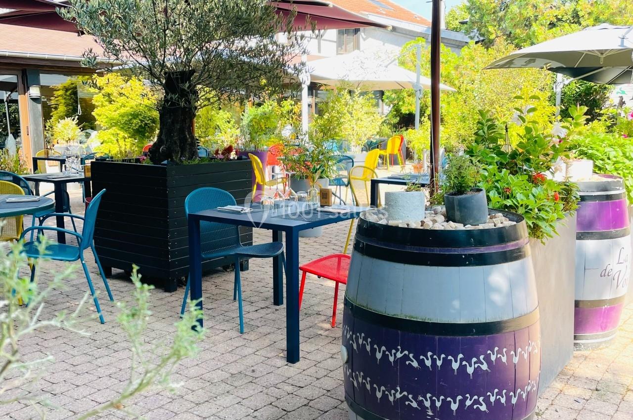 Terrasse extérieure avec tables, chaises colorées, parasols, tonneaux décoratifs et végétation environnante.