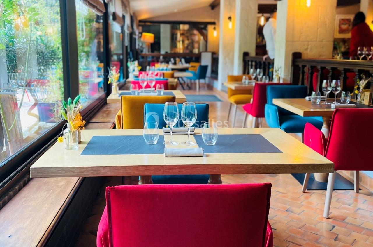 Salle de restaurant lumineuse avec tables dressées, chaises colorées et grandes fenêtres donnant sur un jardin.