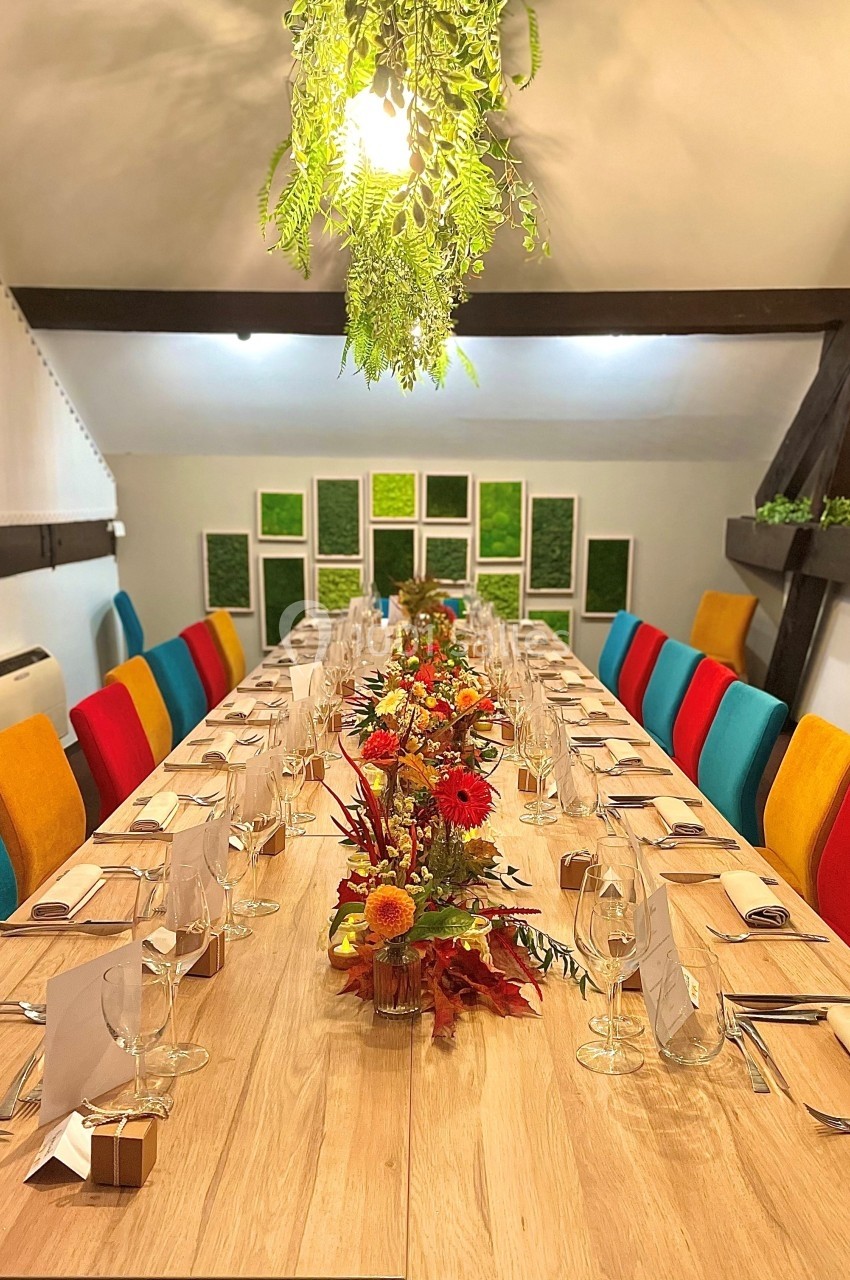 Table en bois dressée pour un repas, décorée de fleurs automnales, entourée de chaises colorées dans une salle lumineuse.