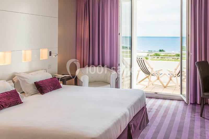 Chambre lumineuse avec lit double, fauteuil, rideaux violets et balcon offrant une vue sur la mer.