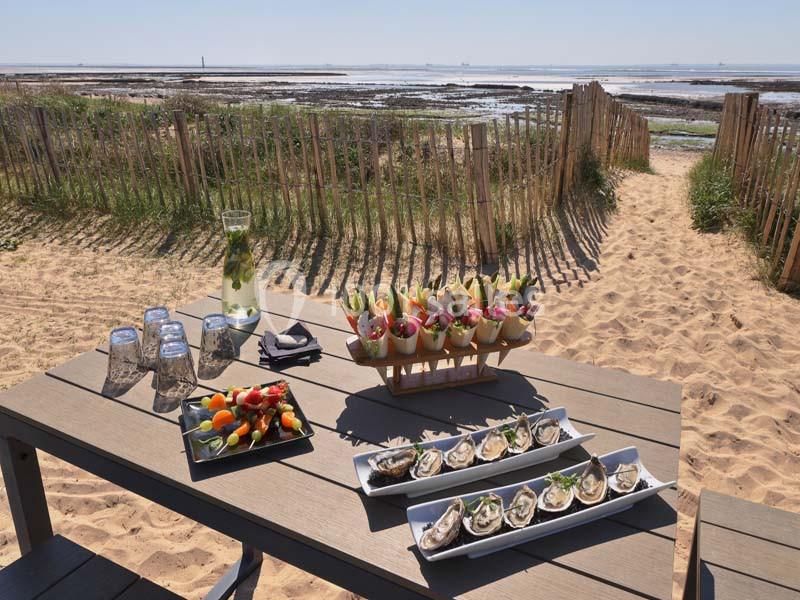 Table en bois sur une plage avec fruits de mer, légumes, boissons et vue sur la mer à travers une clôture en bois.