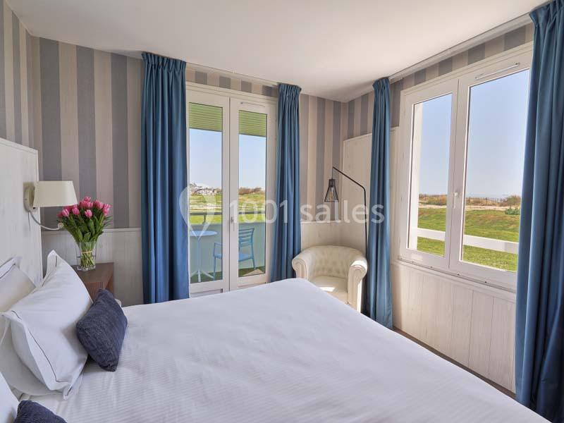 Chambre lumineuse avec lit double, fauteuil, bouquet de fleurs, et vue sur un jardin à travers de grandes fenêtres.