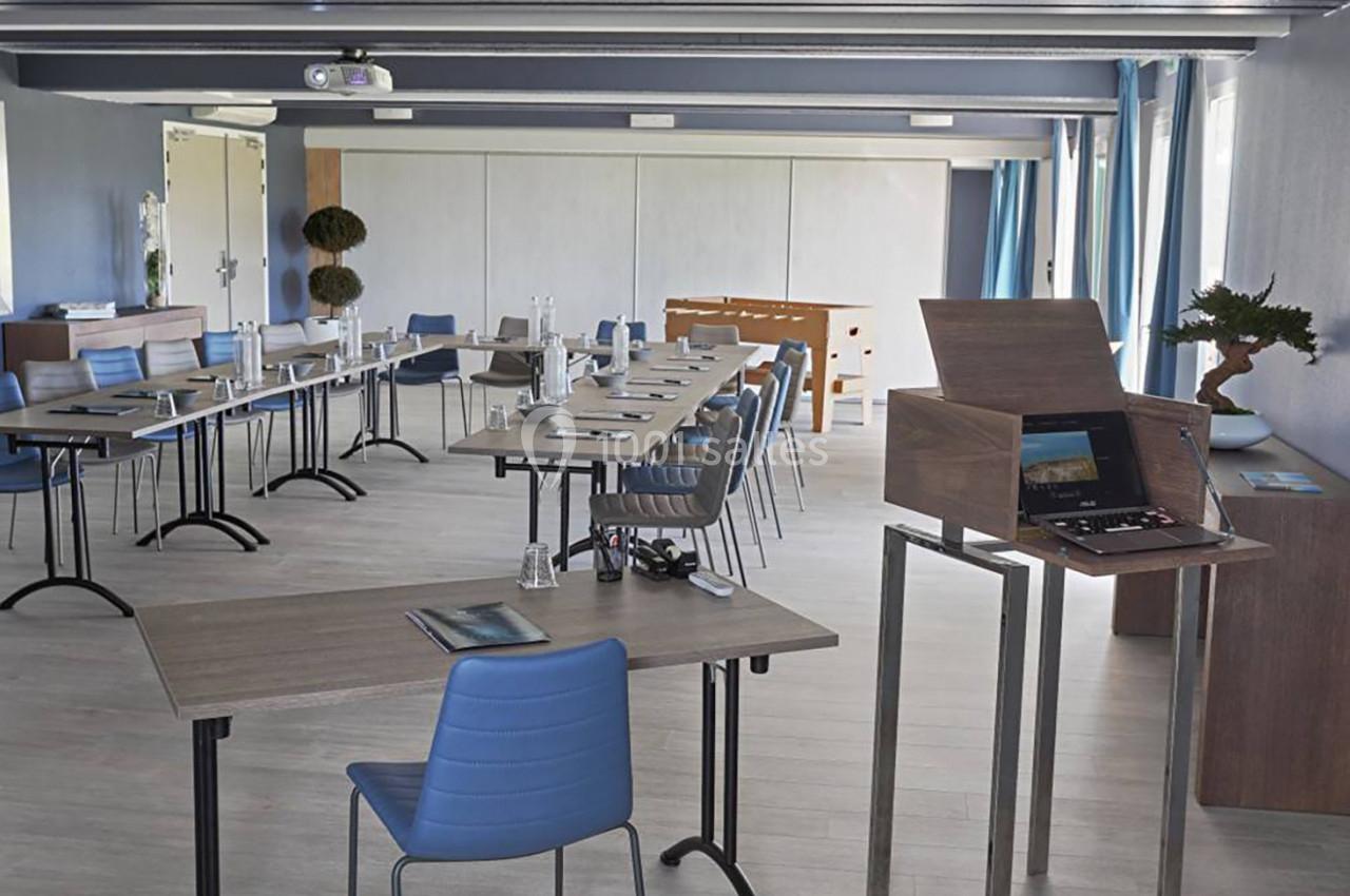 Salle de réunion lumineuse avec tables en bois, chaises bleues, matériel de présentation et grandes fenêtres.