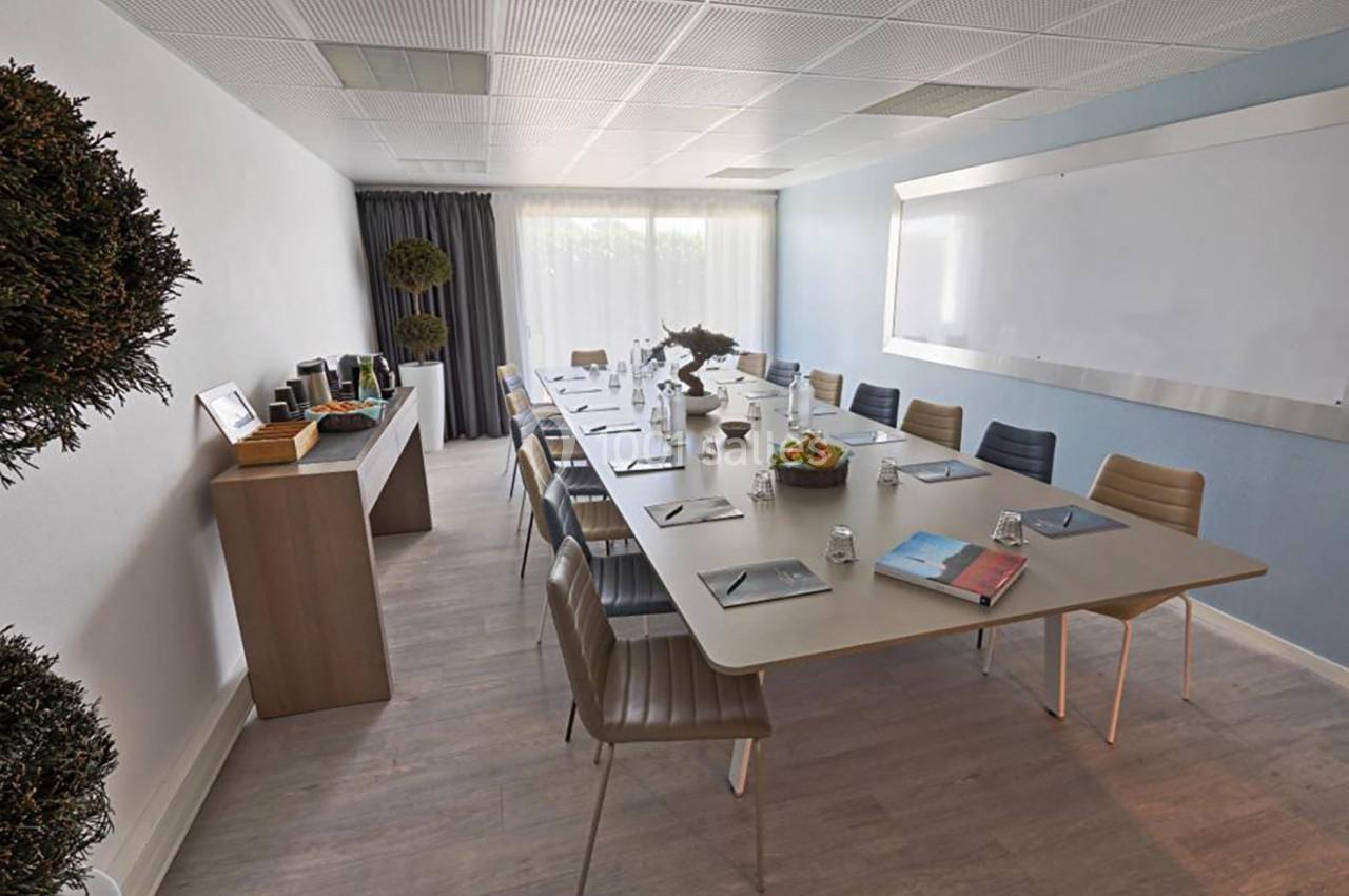 Salle de réunion lumineuse avec une grande table rectangulaire, des chaises, un tableau blanc et un buffet à boissons.