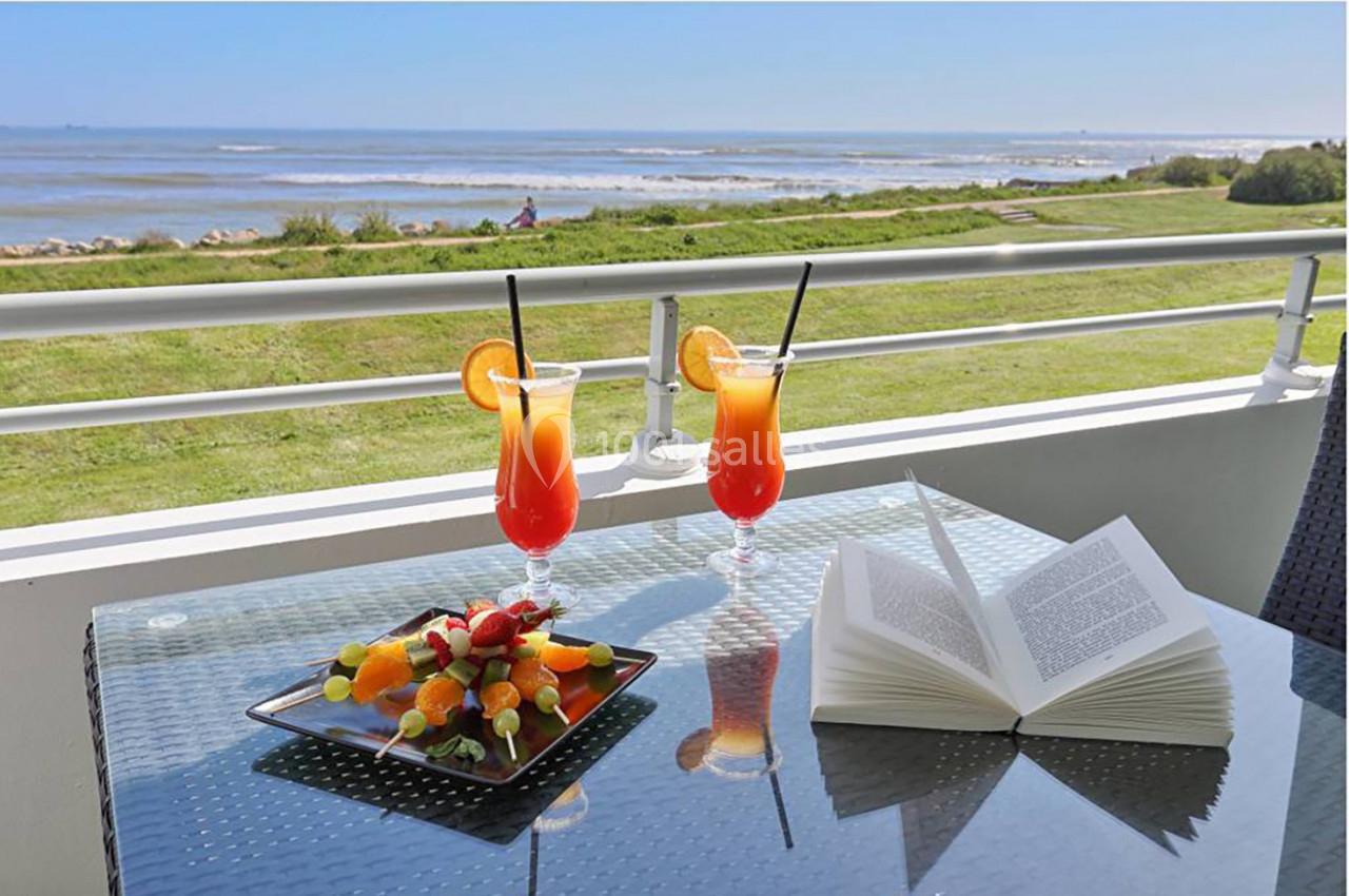 Deux cocktails colorés, une assiette de fruits et un livre ouverts sur une table en terrasse avec vue sur la mer.
