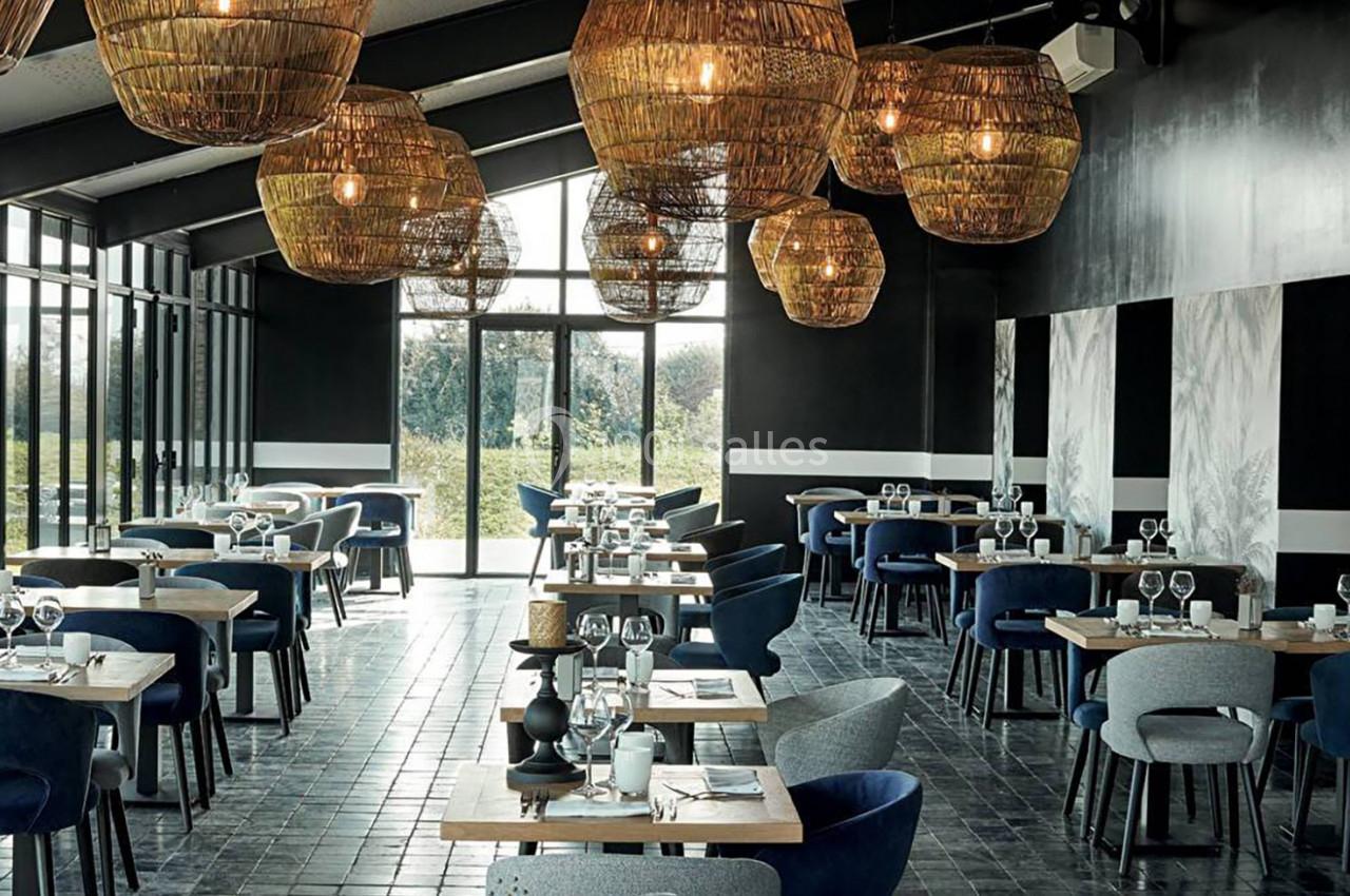 Salle de restaurant moderne avec tables dressées, chaises bleues et grises, grandes baies vitrées et suspensions en osier.