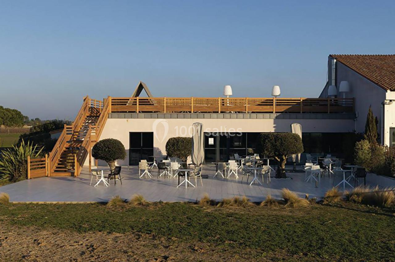 Terrasse extérieure d'un bâtiment avec tables, chaises, parasols et escaliers en bois, entourée de végétation.