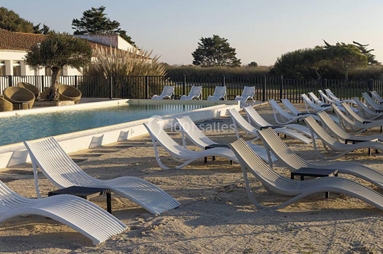Chaises longues blanches alignées autour d'une piscine extérieure, avec un bâtiment et des arbres en arrière-plan.
