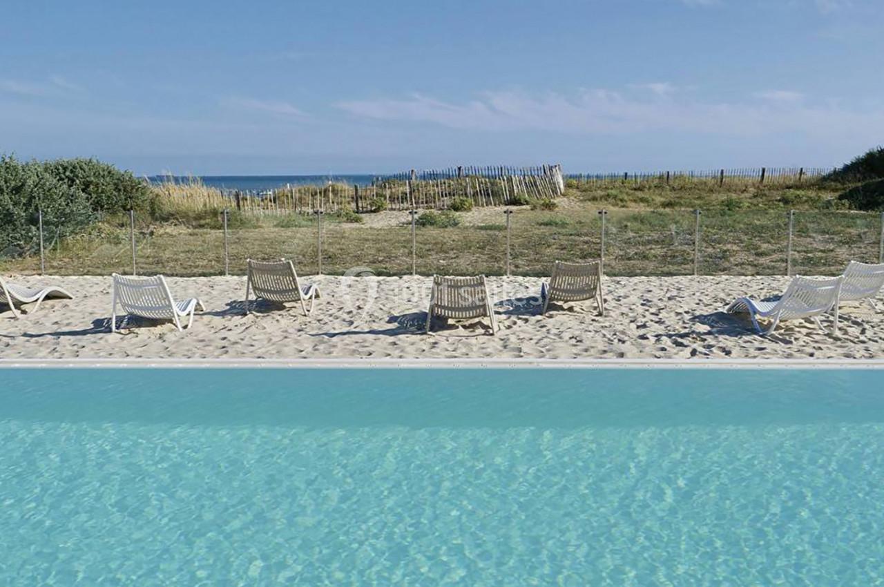 Chaises longues alignées sur du sable près d'une piscine, avec une vue sur une plage et la mer à l'arrière-plan.