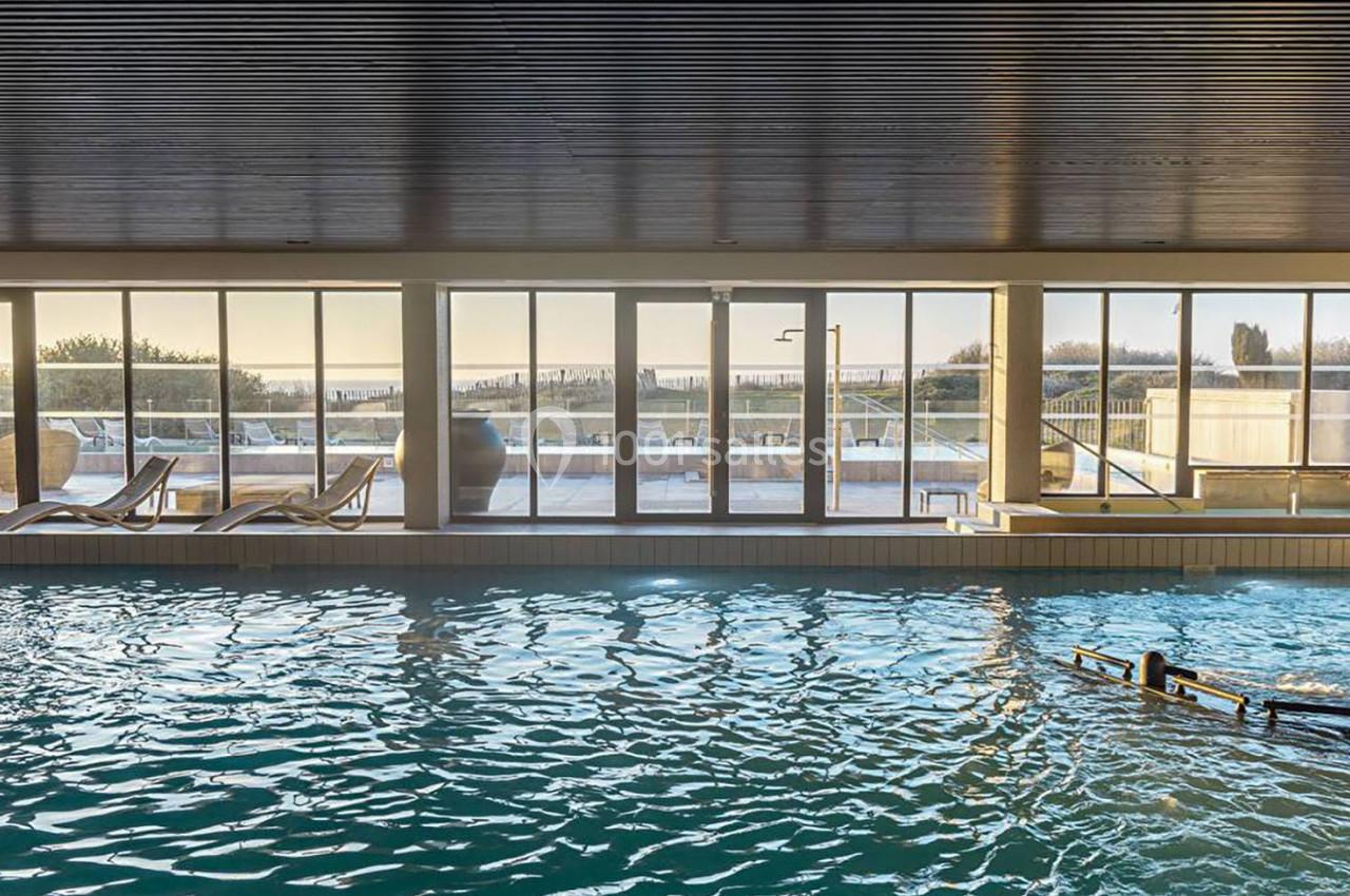 Piscine intérieure avec grandes baies vitrées donnant sur une terrasse ensoleillée et un paysage extérieur.