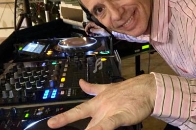 Un DJ souriant en chemise rayée utilise une table de mixage avec des platines et des équipements audio.