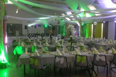 Salle de réception décorée avec des tables dressées, nappes grises, chaises ornées de nœuds verts et éclairage tamisé.