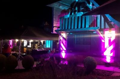 Maison en bois éclairée par des lumières colorées la nuit, avec un stand et du matériel technique à l'extérieur.