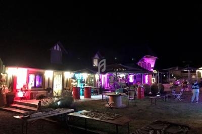 Bâtiment éclairé par des lumières colorées la nuit, avec des tables et parasols disposés sur une pelouse.