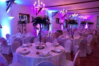 Salle de réception décorée pour un mariage, avec tables rondes dressées, chaises blanches et éclairage rose.