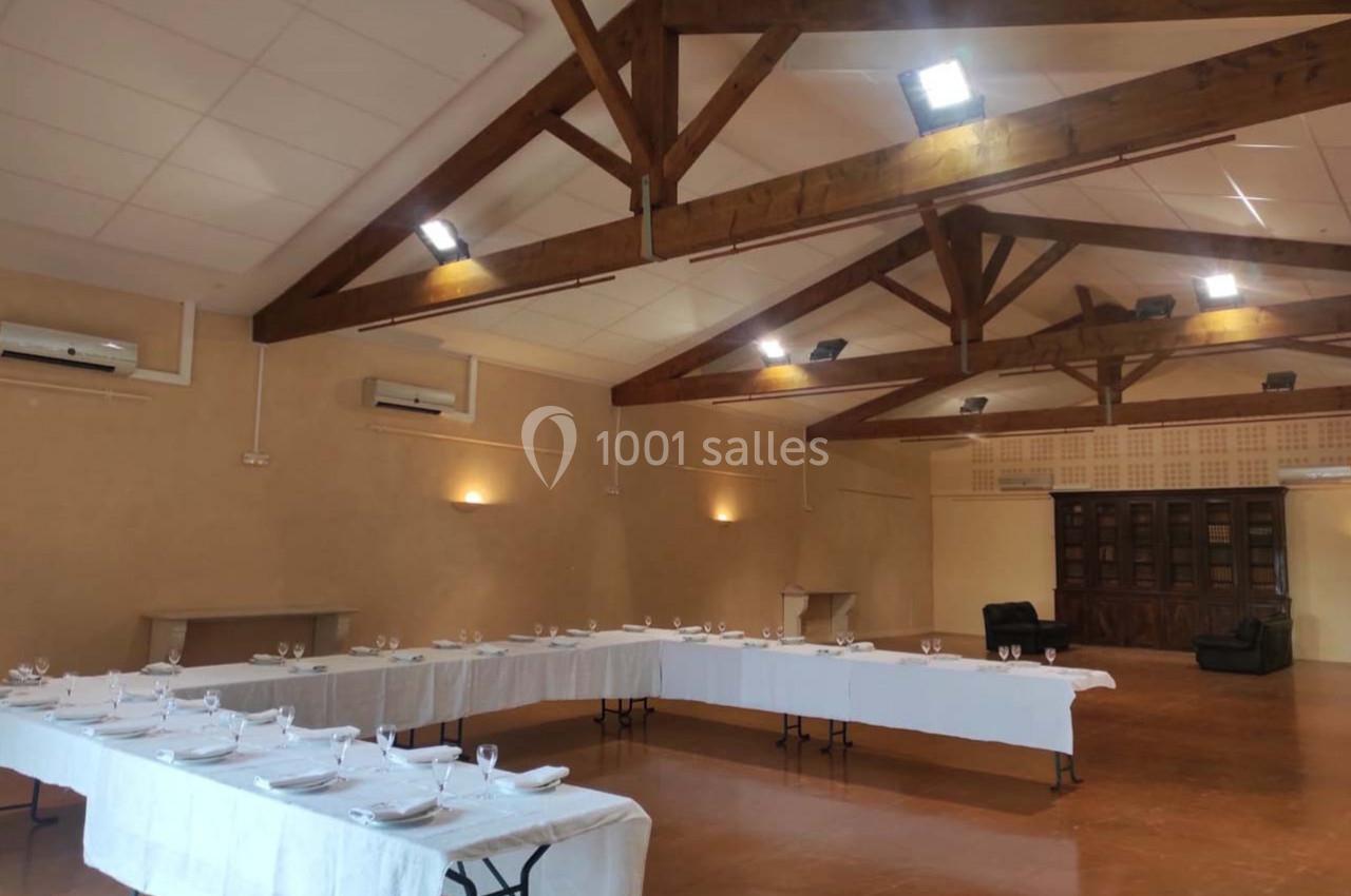 Salle spacieuse avec tables dressées en U, nappes blanches, vaisselle et poutres apparentes au plafond.