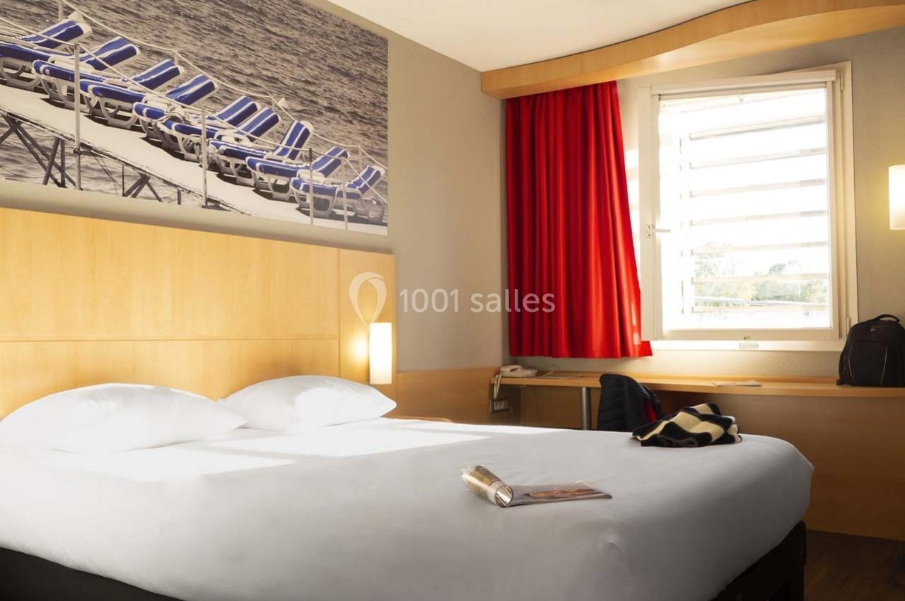Chambre d'hôtel lumineuse avec un lit double, une fenêtre avec rideaux rouges et un bureau en bois clair.