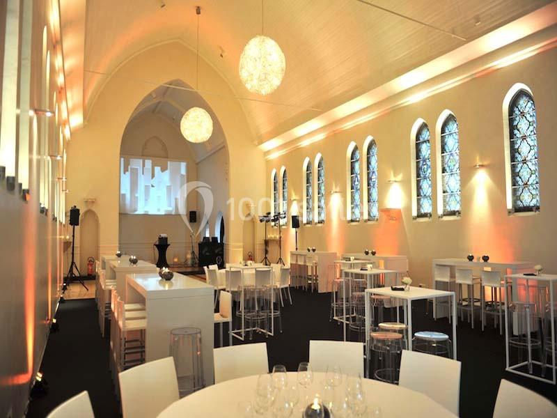 Salle lumineuse avec hauts plafonds voûtés, grandes fenêtres en ogive et mobilier moderne blanc.