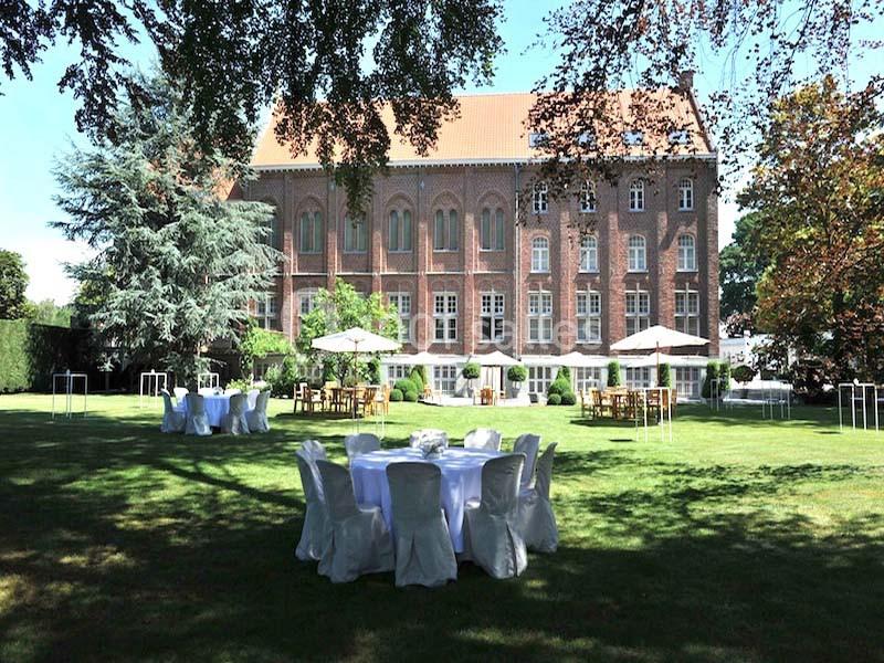 Jardin avec tables rondes dressées sous des parasols, devant un grand bâtiment en briques rouges.
