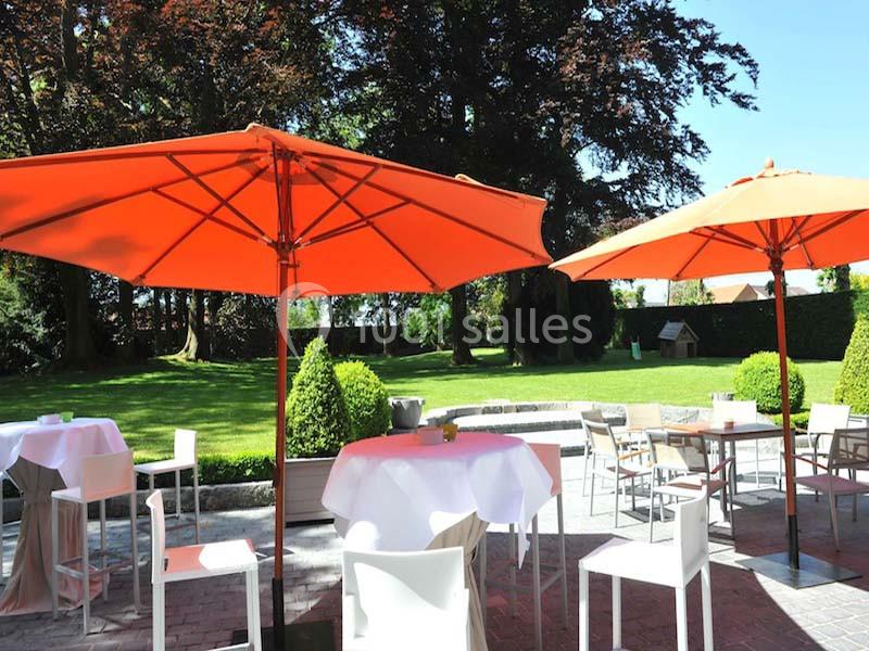 Terrasse avec tables et chaises blanches sous de grands parasols orange, donnant sur un jardin verdoyant.