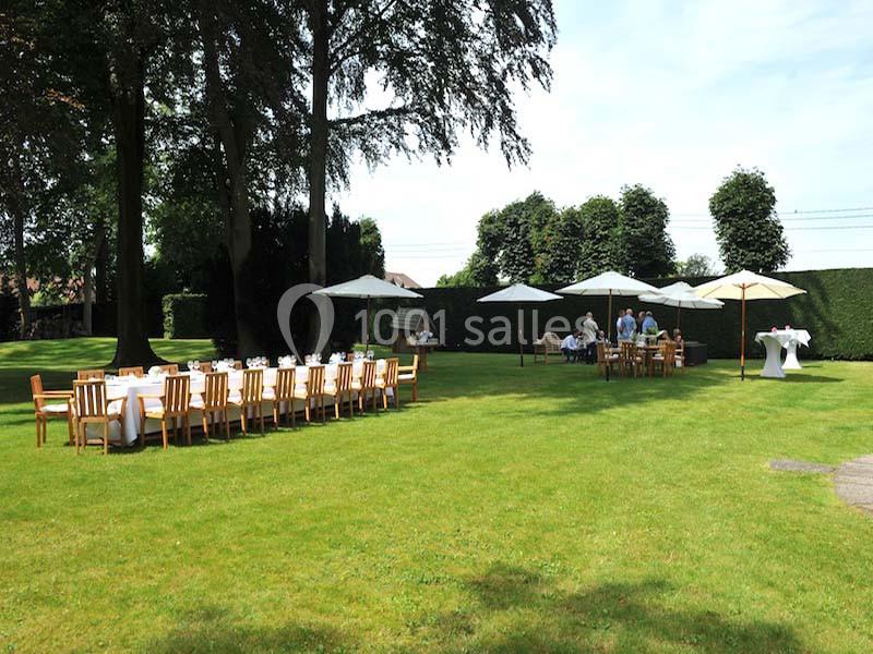 Tables et chaises en bois disposées sur une pelouse, avec parasols et personnes en arrière-plan dans un jardin.