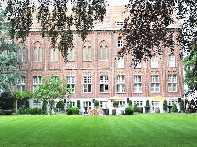 Façade en briques rouges d'un bâtiment historique entouré d'arbres, avec une pelouse et des tables en terrasse.