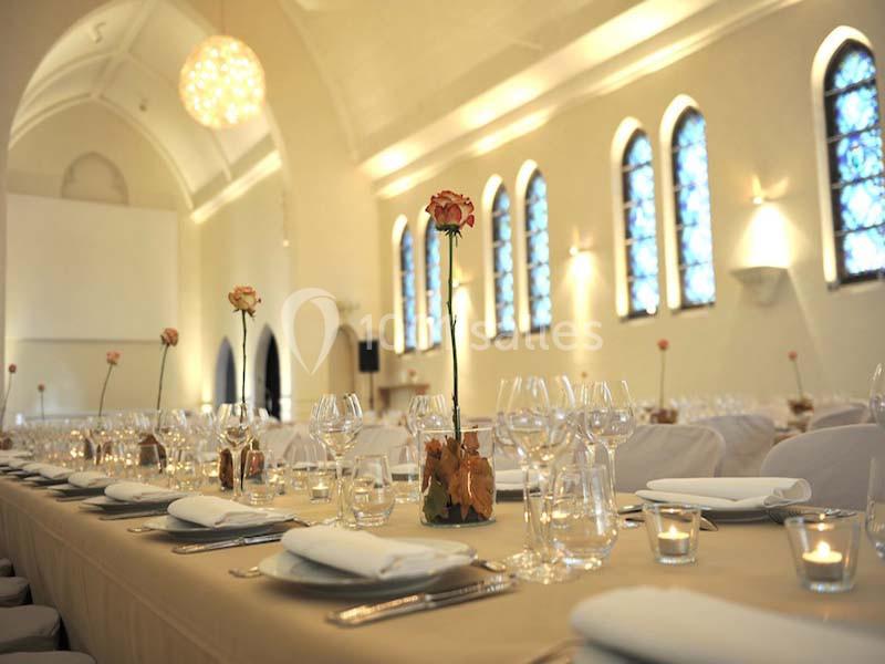 Salle de réception élégante avec longues tables dressées, décorées de roses et éclairées par des vitraux colorés.