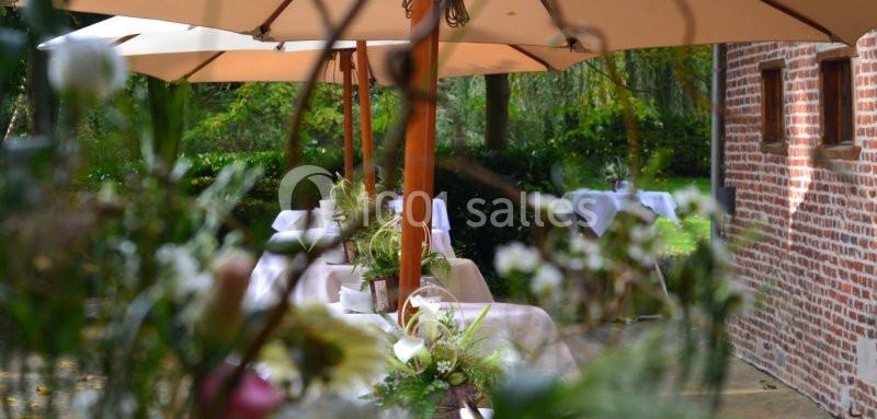 Terrasse extérieure avec tables dressées sous des parasols, entourée de verdure et d'un mur en briques rouges.