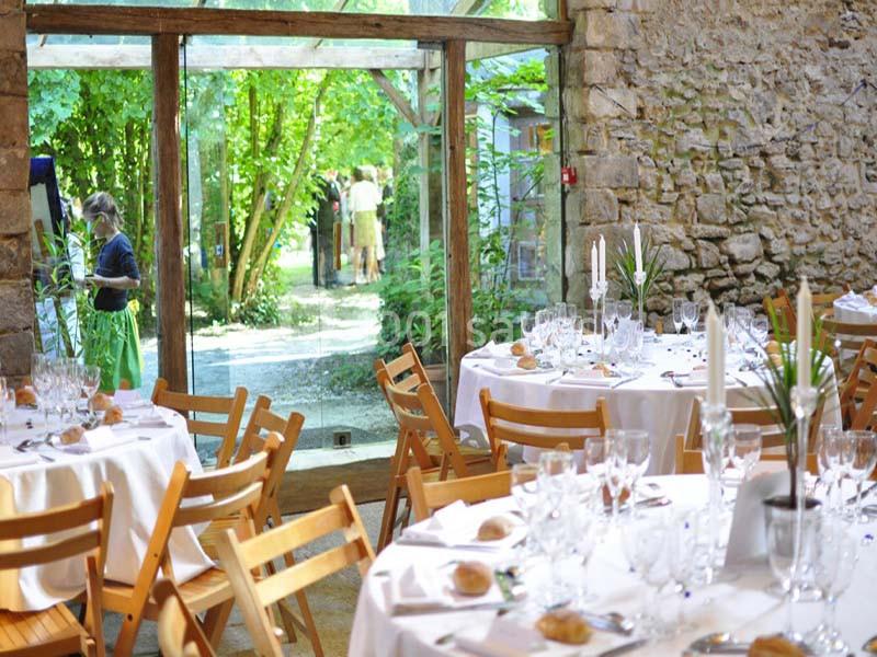 Salle de réception avec tables dressées, vue sur un jardin verdoyant à travers une large ouverture vitrée.