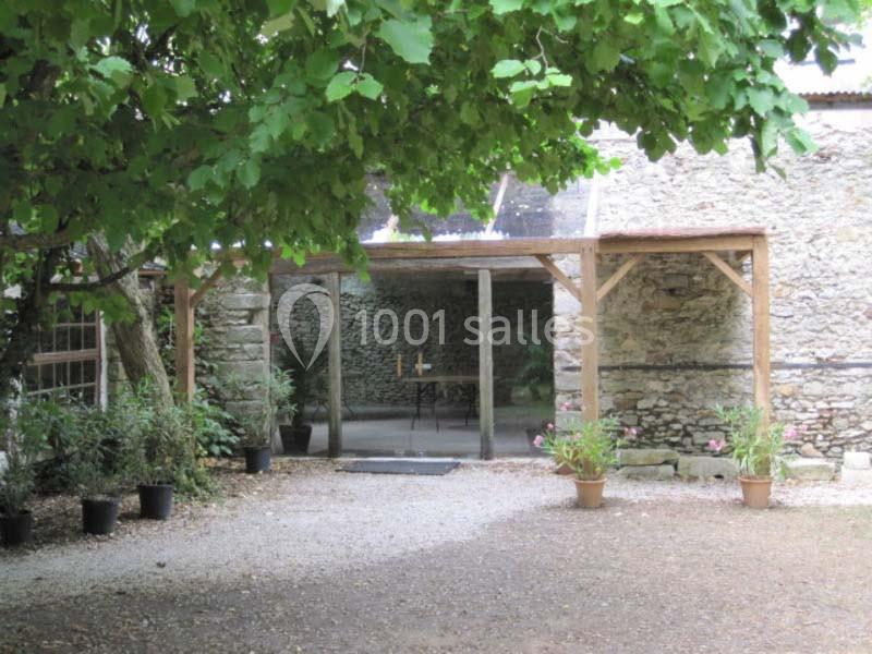 Entrée vitrée d'un bâtiment en pierre entourée de plantes en pots et d'un arbre ombrageant la cour.