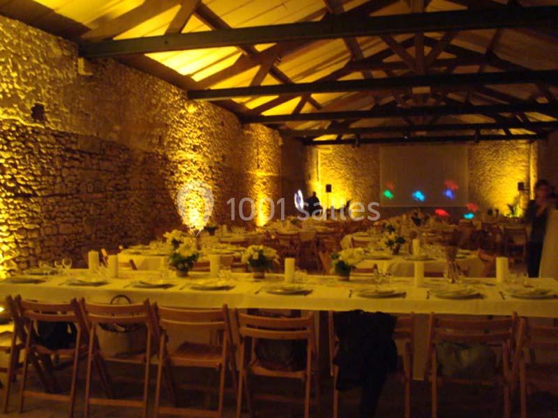 Salle en pierre éclairée de lumières chaudes, avec de longues tables dressées pour un repas festif.