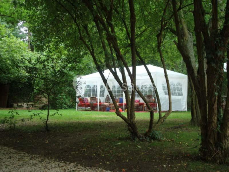 Tente blanche installée dans un jardin verdoyant, entourée d'arbres et de mobilier visible à l'intérieur.