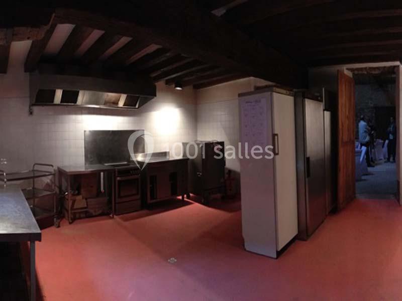 Cuisine professionnelle avec sol rouge, équipements en inox, réfrigérateurs et hotte sous un plafond en poutres apparentes.