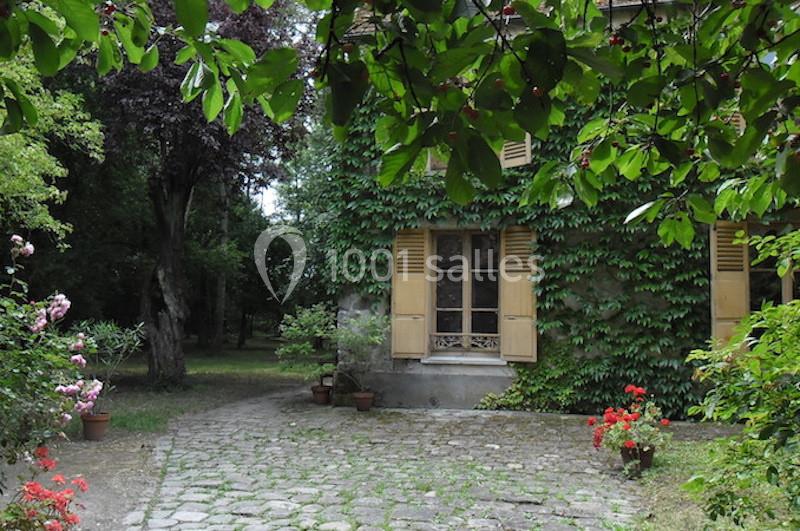 Façade d'une maison recouverte de lierre, avec des volets en bois et une cour pavée entourée de végétation.