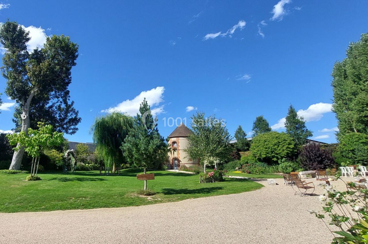 Jardin verdoyant avec pelouse, arbres, allée gravillonnée et bâtiment en briques au toit conique sous un ciel bleu.