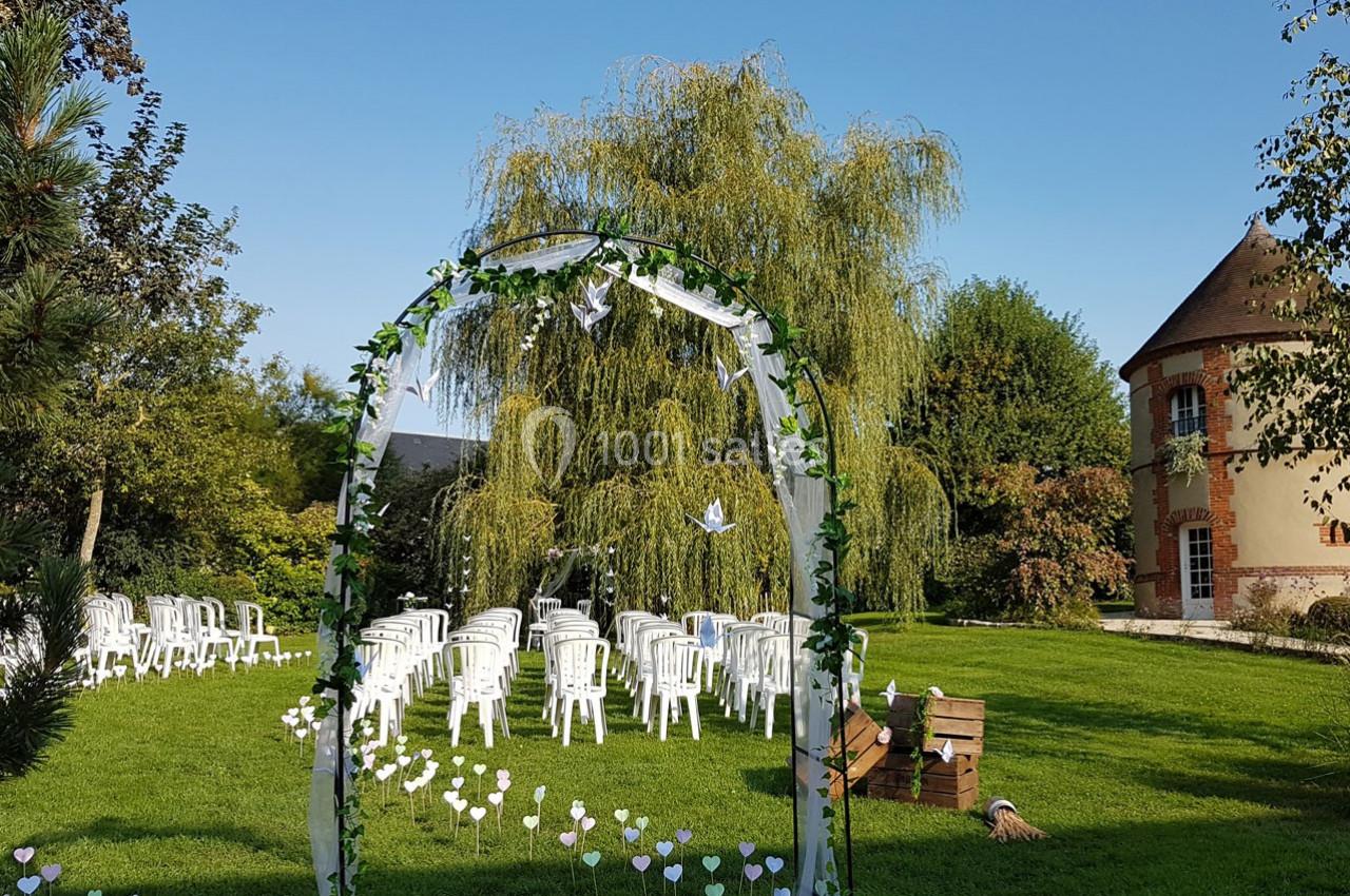 Arche décorée de feuillage et de colombes en papier, entourée de chaises blanches disposées sur une pelouse devant un saule…