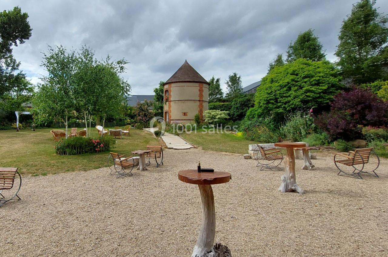 Jardin avec bancs, tables en bois et un pigeonnier en arrière-plan sous un ciel nuageux.