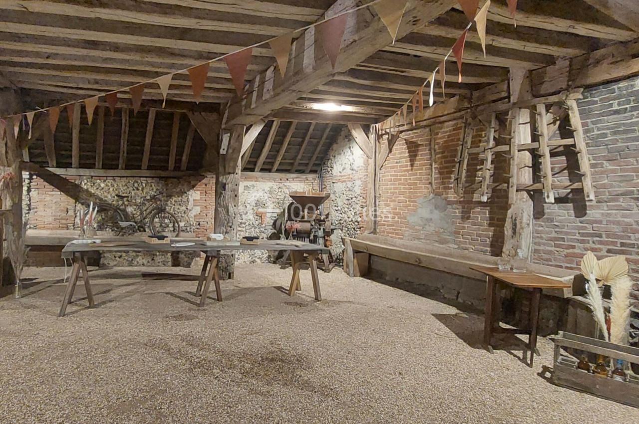 Intérieur d'une grange rustique avec poutres apparentes, guirlandes, outils agricoles et tables en bois.