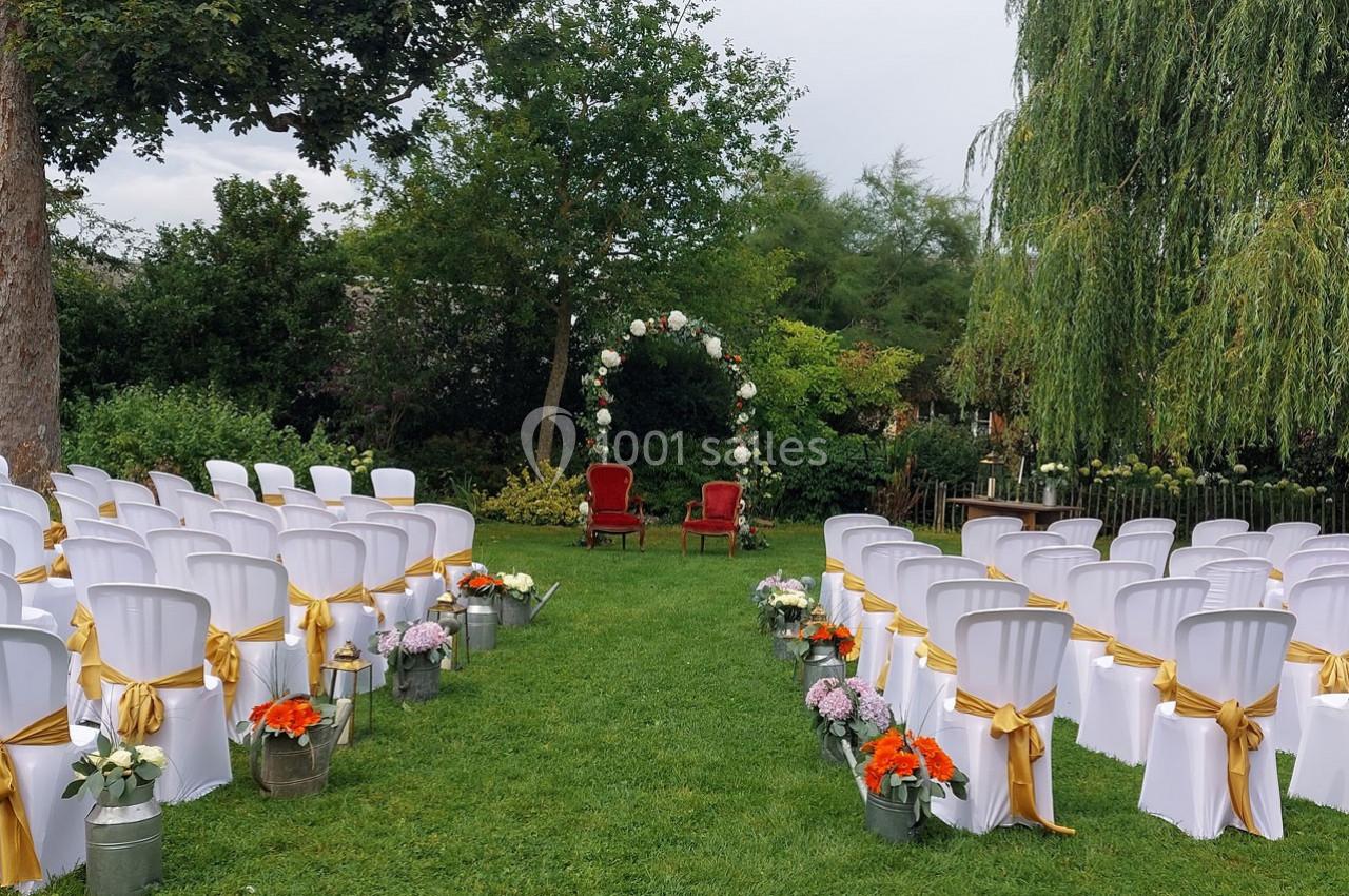 Chaises blanches décorées de rubans dorés disposées en allée, avec deux fauteuils rouges sous une arche fleurie dans un…