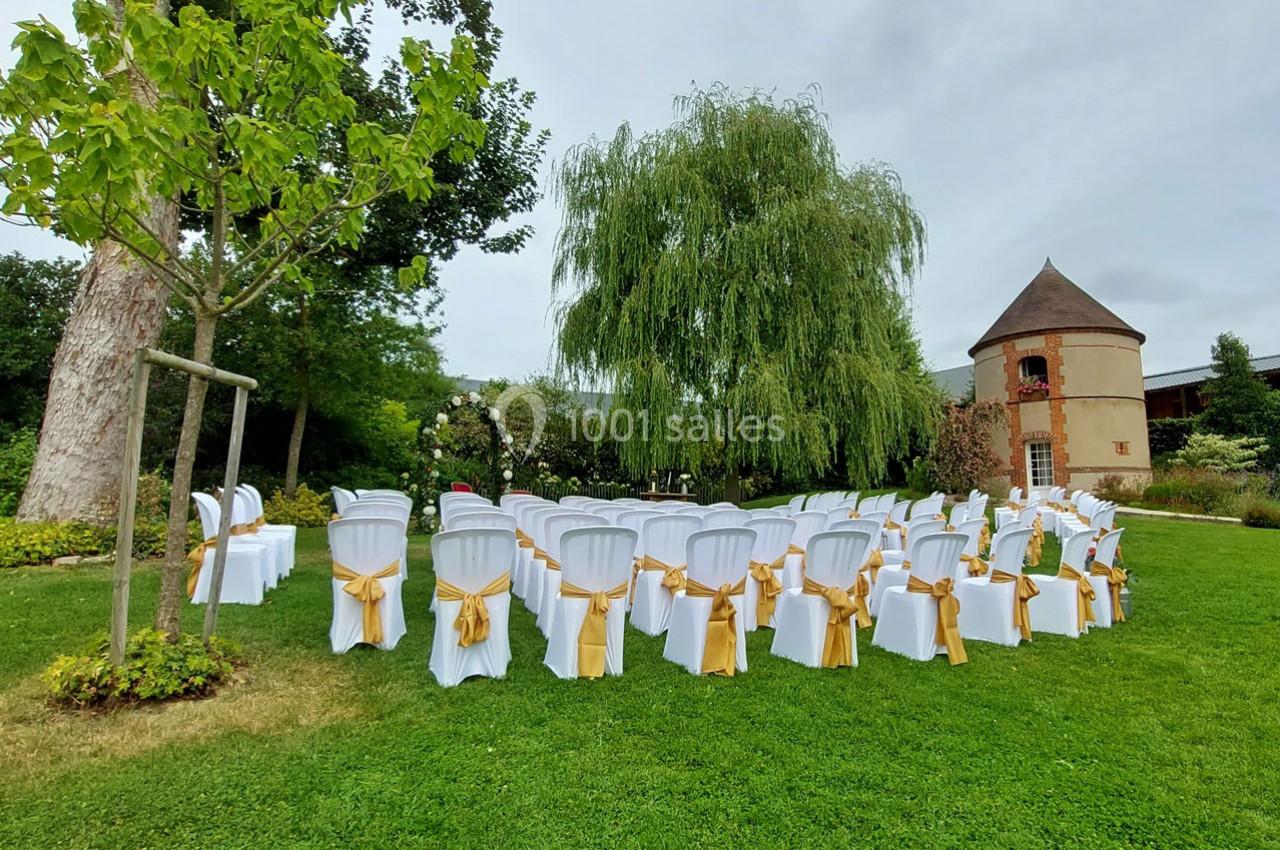 Chaises blanches décorées de rubans dorés disposées en extérieur devant un arbre et un bâtiment en pierre.