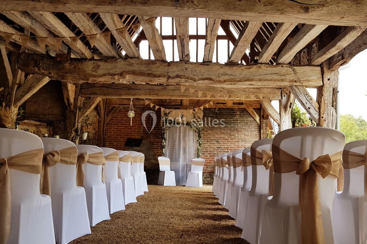 Salle de réception rustique avec des chaises blanches décorées de nœuds en toile de jute, alignées face à une arche fleurie.