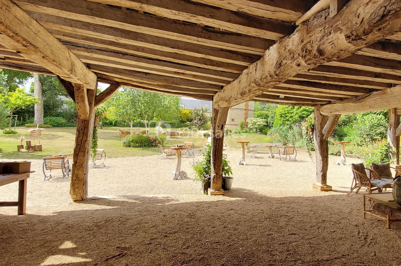 Vue d'un espace extérieur avec une structure en bois, donnant sur un jardin verdoyant et des tables disposées.