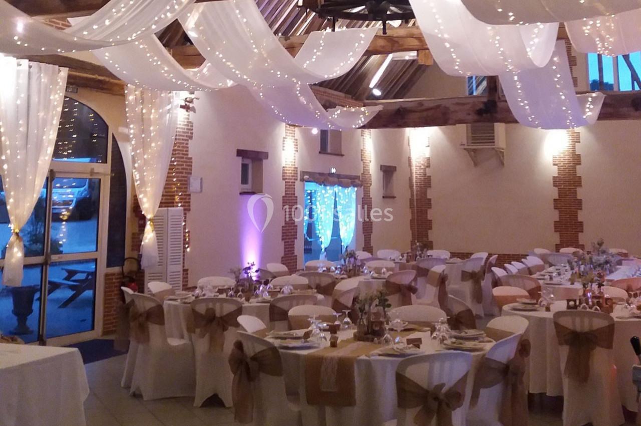Salle de réception décorée pour un mariage avec tables dressées, chaises ornées de nœuds et guirlandes lumineuses suspendues.