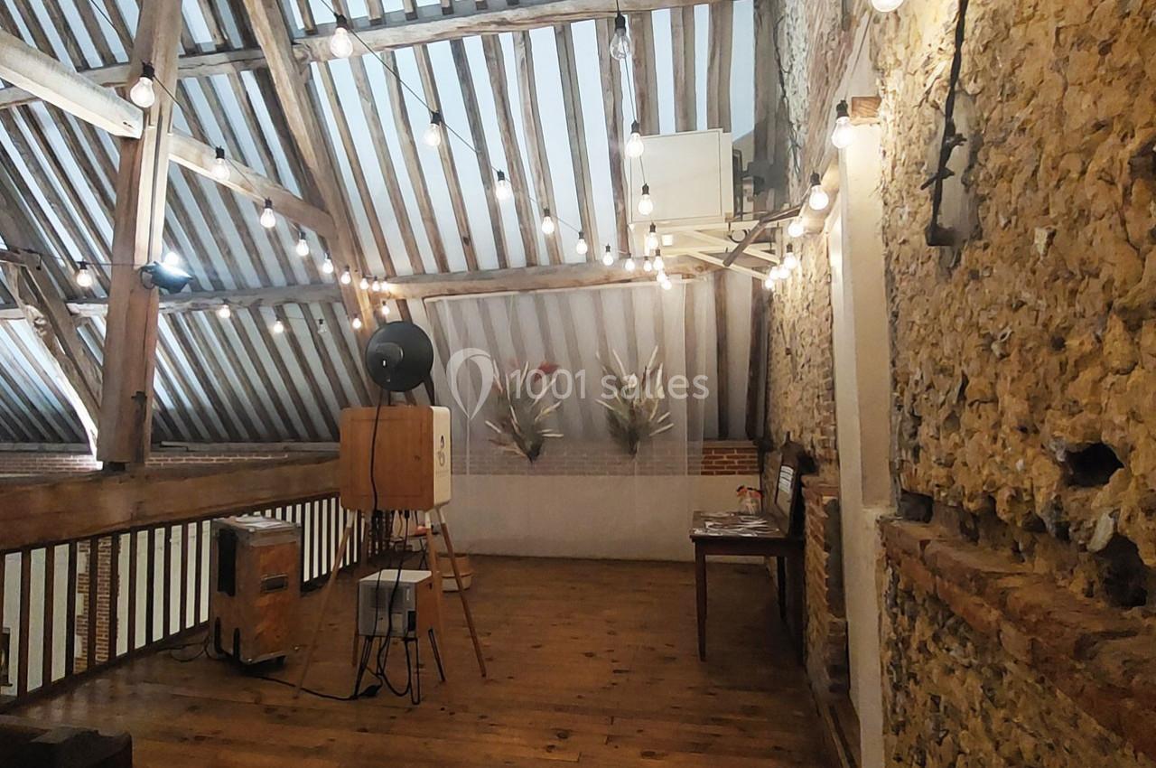 Grenier aménagé avec poutres apparentes, murs en pierre, guirlandes lumineuses et mobilier en bois.