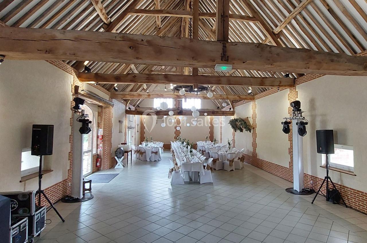 Salle de réception avec charpente en bois apparente, tables dressées pour un événement et éclairage installé.